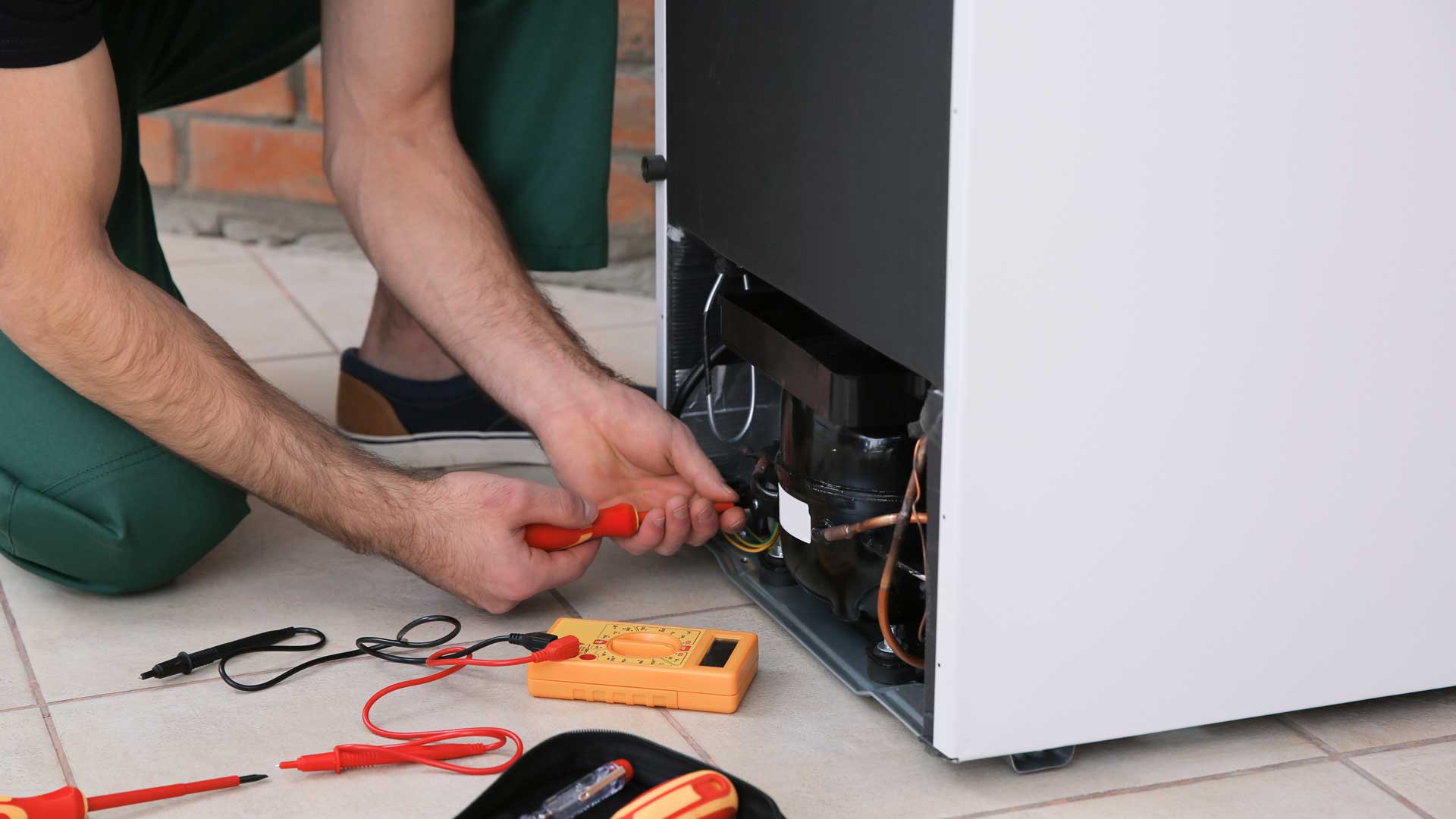 A man kneeling behind a refrigerator to tinker with the wiring on the back